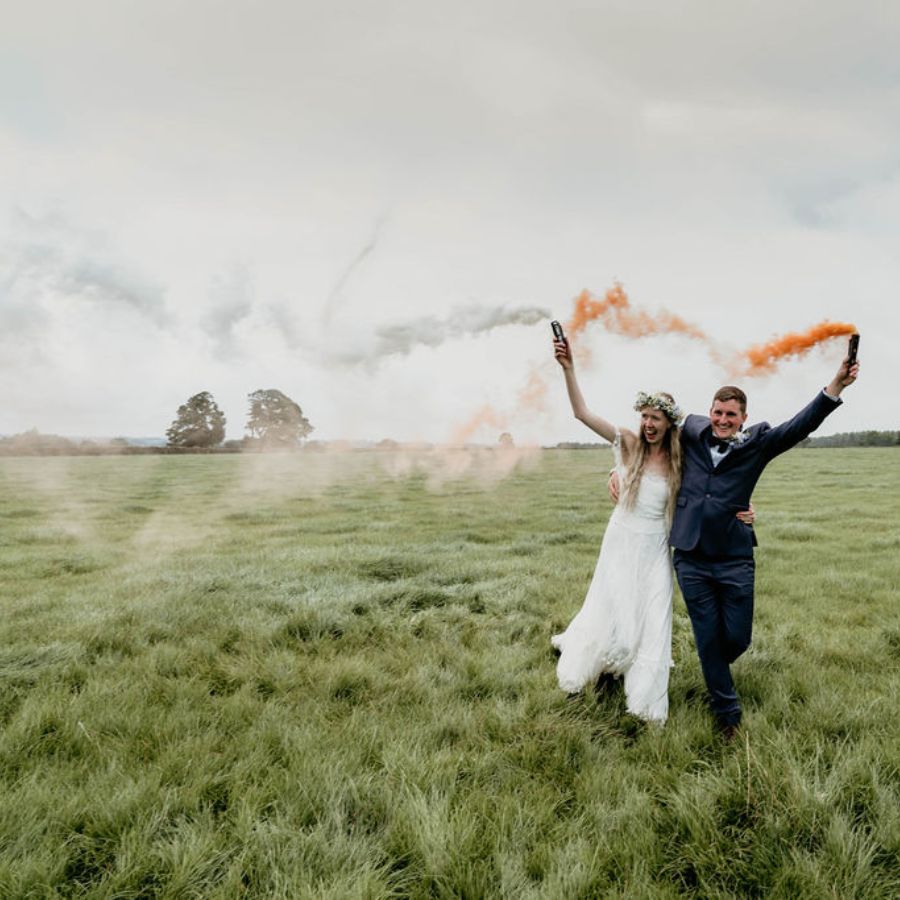 Newly married couple run through field with colourful smoke flairs